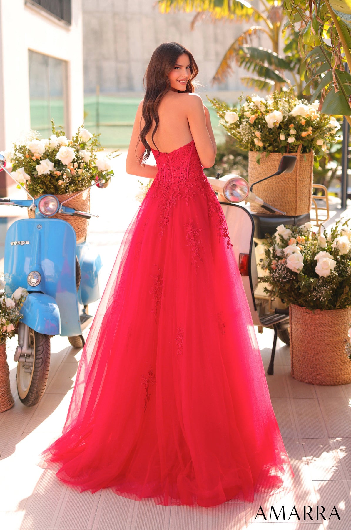 Woman in a red dress standing outdoors with flowers and a scooter in the background, brand 'AMARRA' visible. THE BRIDAL COMPANY