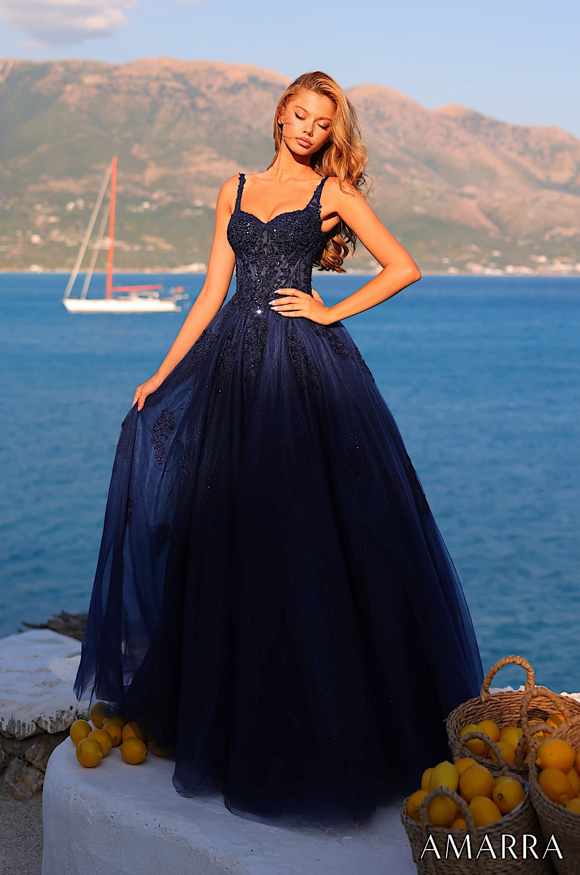 Woman in a navy blue evening gown standing by the sea with mountains in the background, brand 'AMARRA' visible.