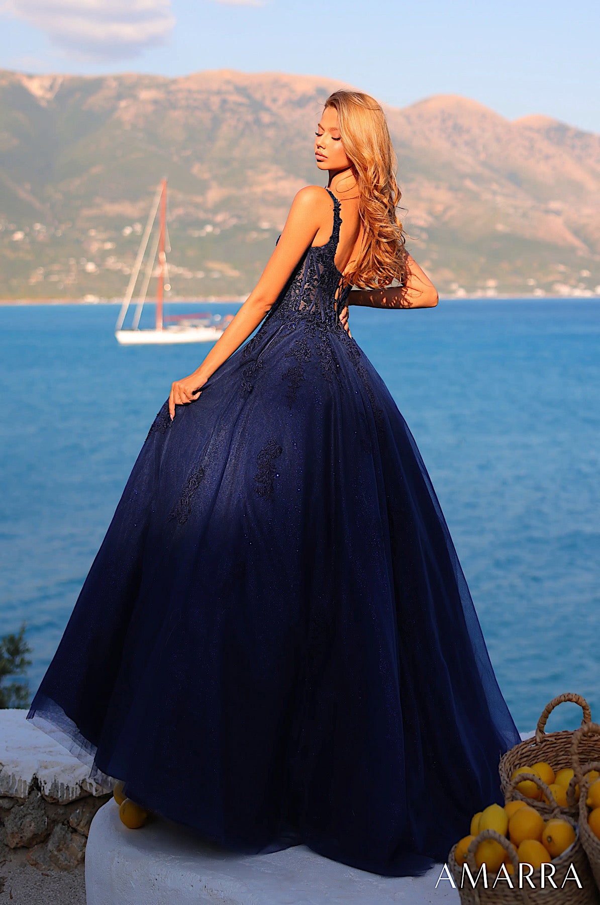 Woman in a navy blue dress standing by the sea with mountains in the background, wearing an 'AMARRA' dress.