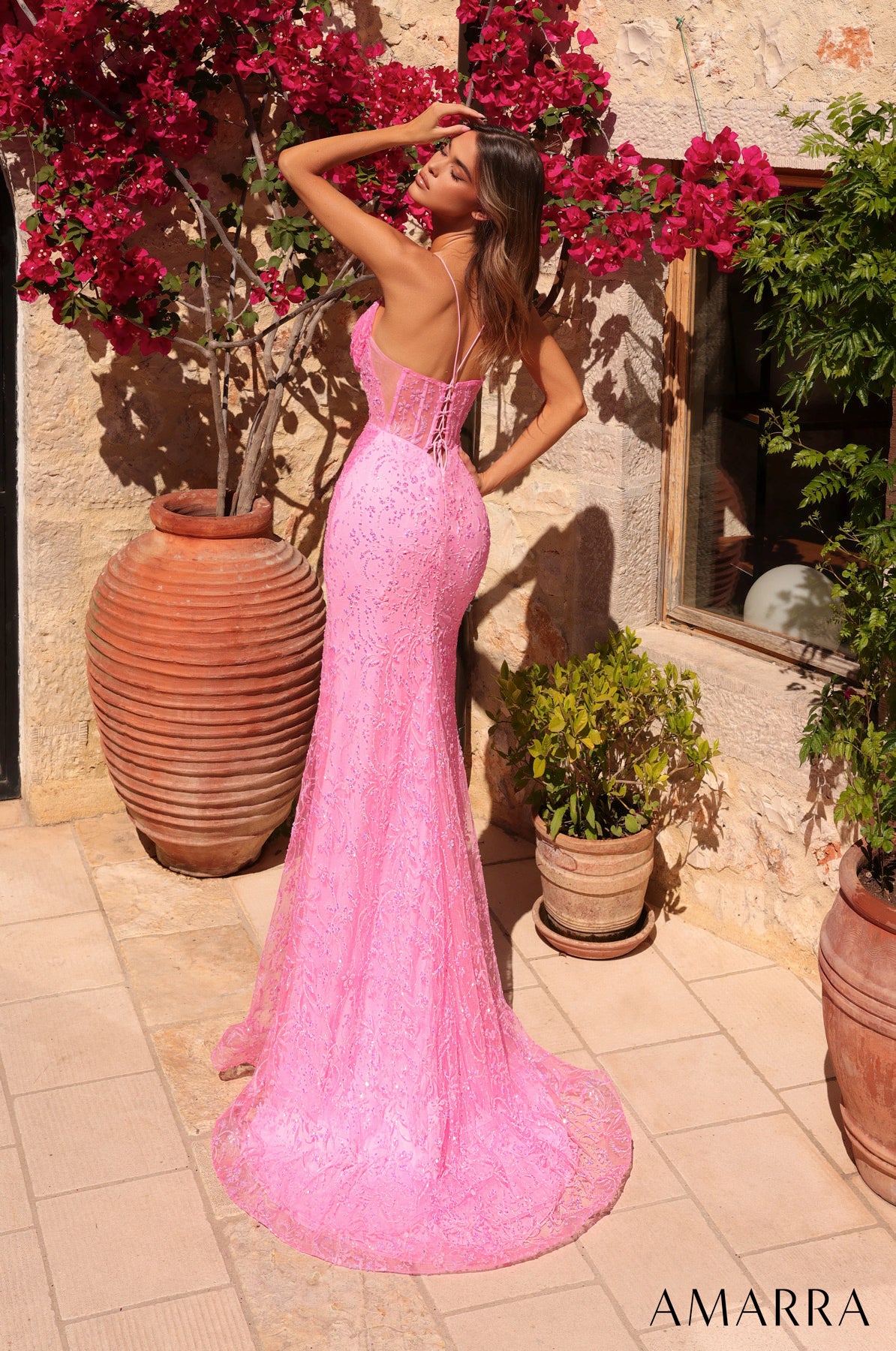 Woman in a pink lace dress standing outdoors with flowers and plants around.