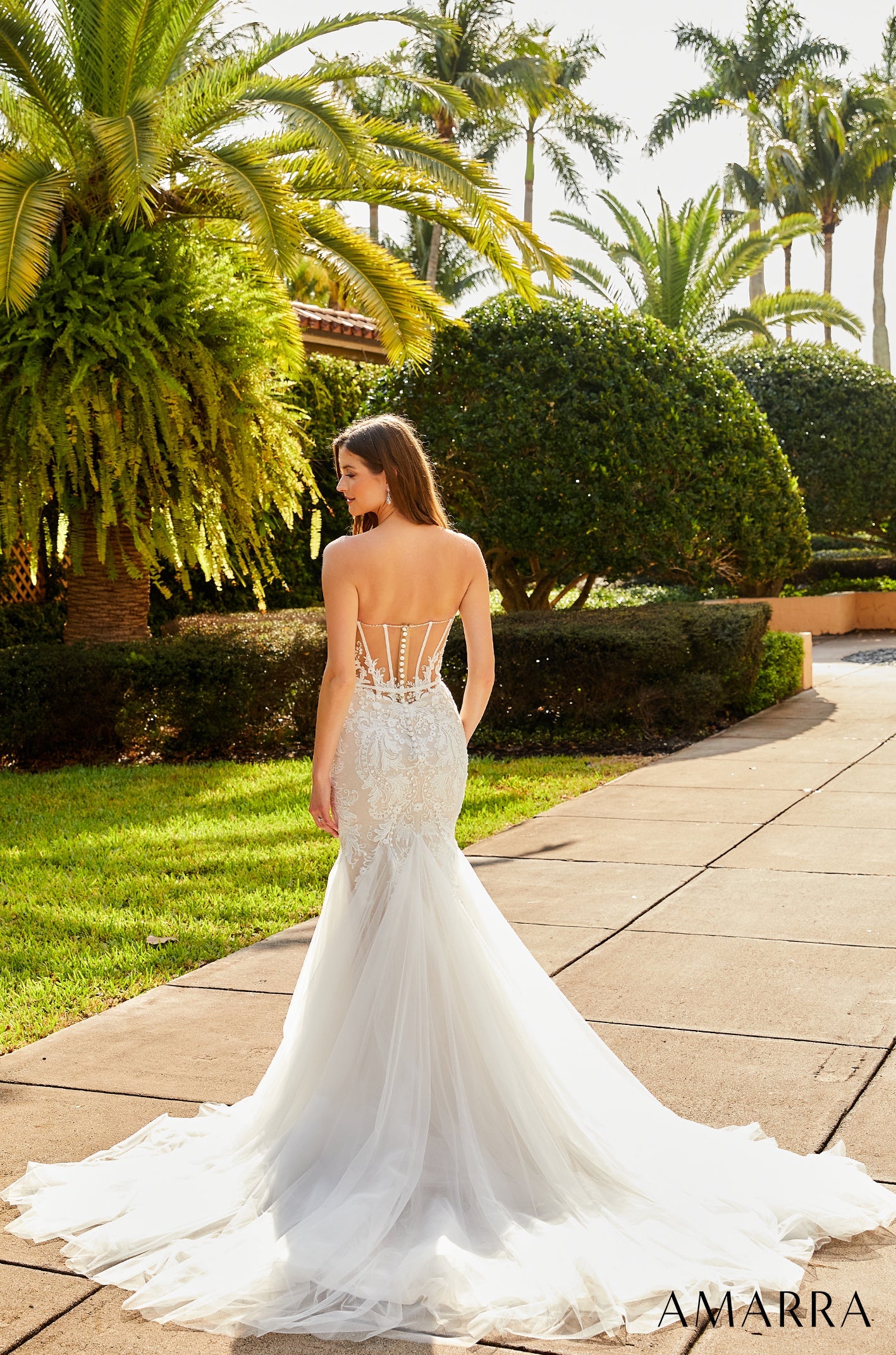 Woman in a white wedding dress with an open back standing on a path with greenery and palm trees in the background.