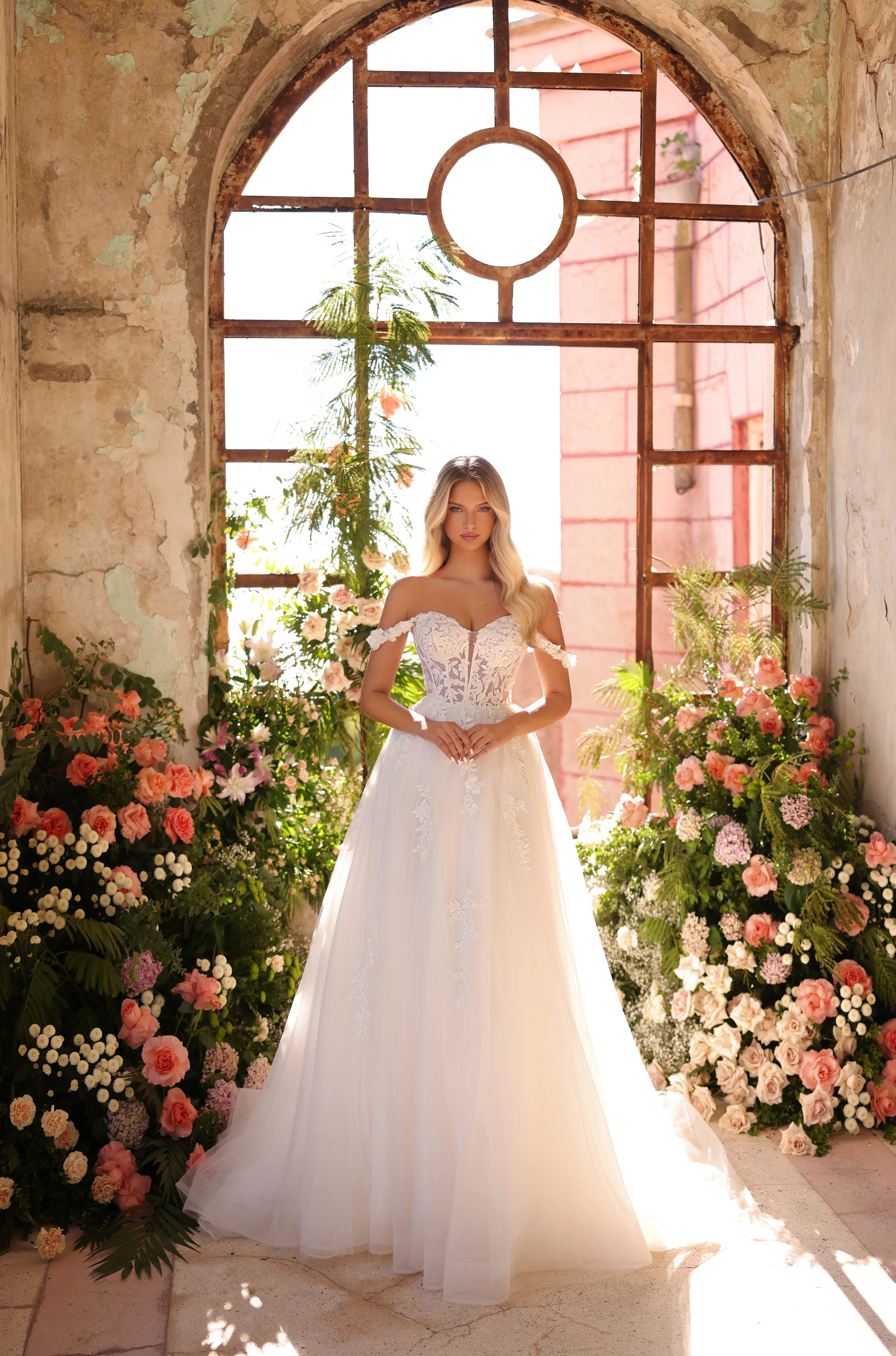 Woman in a wedding dress standing in front of a large window with floral decorations.